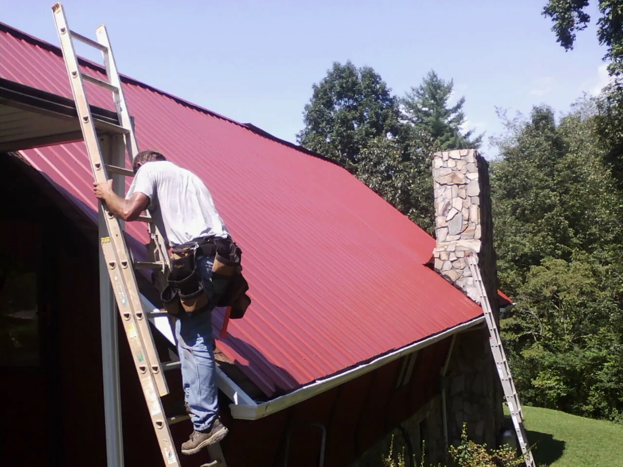 Roofer on a ladder beside a red metal roof and stone chimney — Soffit & Fascia Repair by Roofing Brothers in La Verkin